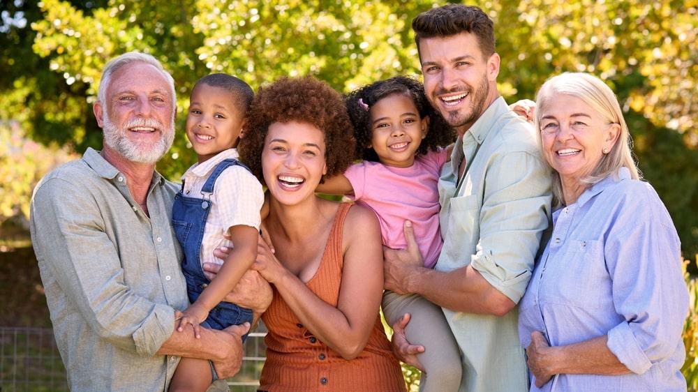 A joyful family of six poses outdoors, including two senior adults, two young adults, and two children. They are smiling in a sunny park setting with trees in the background, all embracing each other warmly.