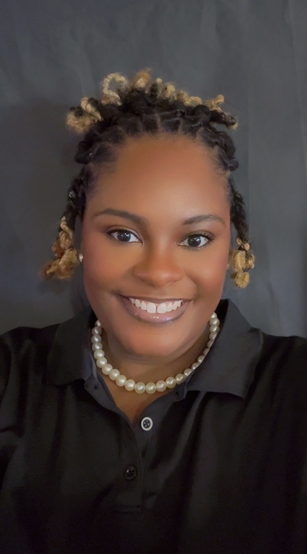 A woman with styled hair in twists, wearing a black collared shirt and a pearl necklace, smiles at the camera against a plain dark background.