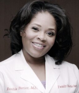 A woman with shoulder-length dark hair smiles, wearing a white lab coat embroidered with Family Medicine and M.D. She stands against a dark background.
